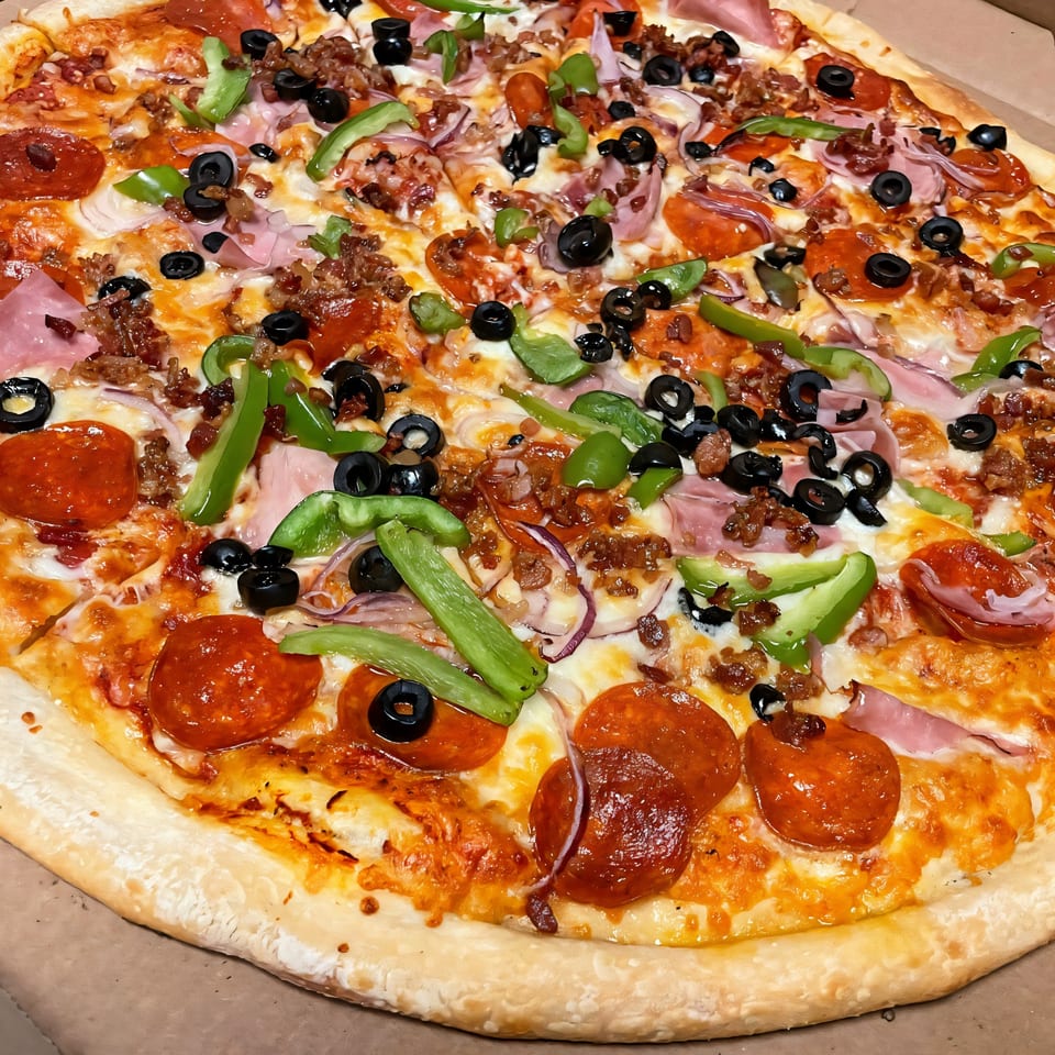 A close-up of a Di Angelo Pizza with pepperoni, ham, green peppers, black olives, and bacon.