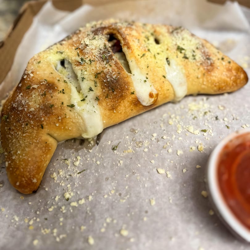 A golden-brown calzone from Di Angelo Pizza, topped with melted cheese, herbs, and parmesan, with a side of marinara.