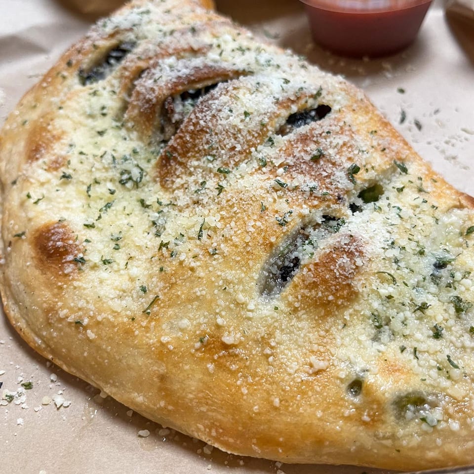 A golden-brown calzone from Di Angelo Pizza, topped with parmesan cheese and herbs, with a side of marinara.